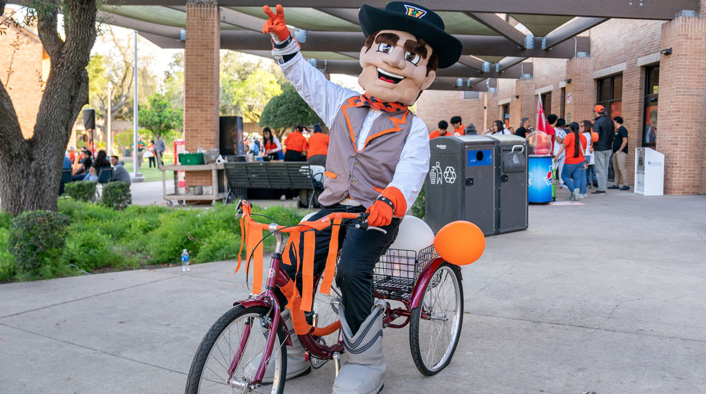 Vaquero mascot riding a bicycle with V's up!