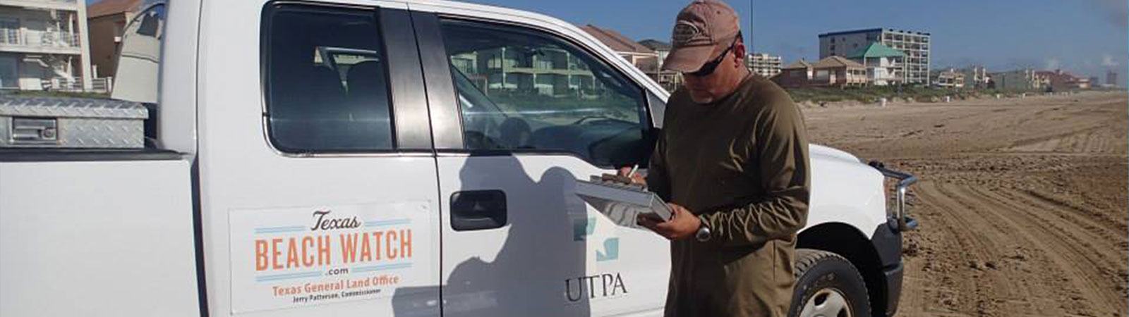 A person stands beside a white pickup truck on a sandy beach. The truck features the “Texas Beach Watch” logo, the Texas General Land Office emblem, and the text “UTPA.” The person wears a cap, sunglasses, and holds a clipboard or tablet. This image likely relates to fieldwork or environmental monitoring by Texas Beach Watch. 