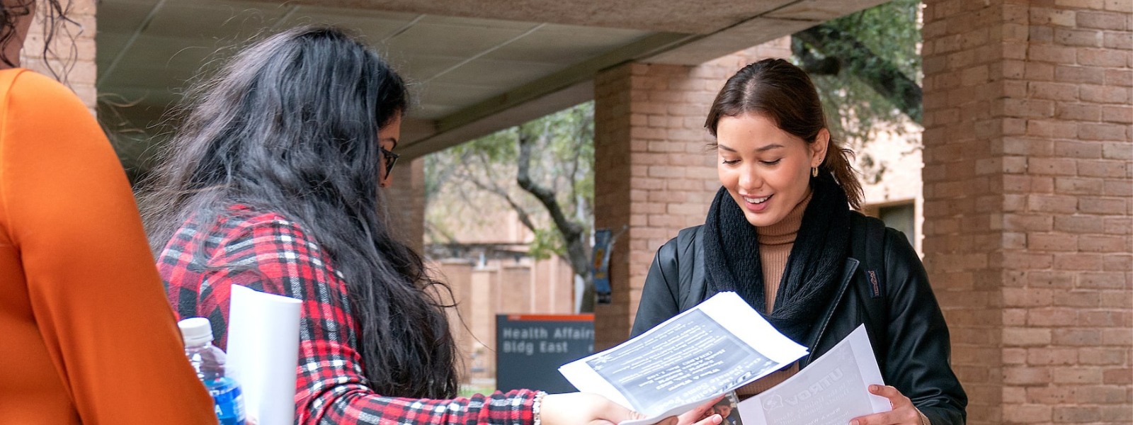 Students smiling, as one student hands the second student a flyer.