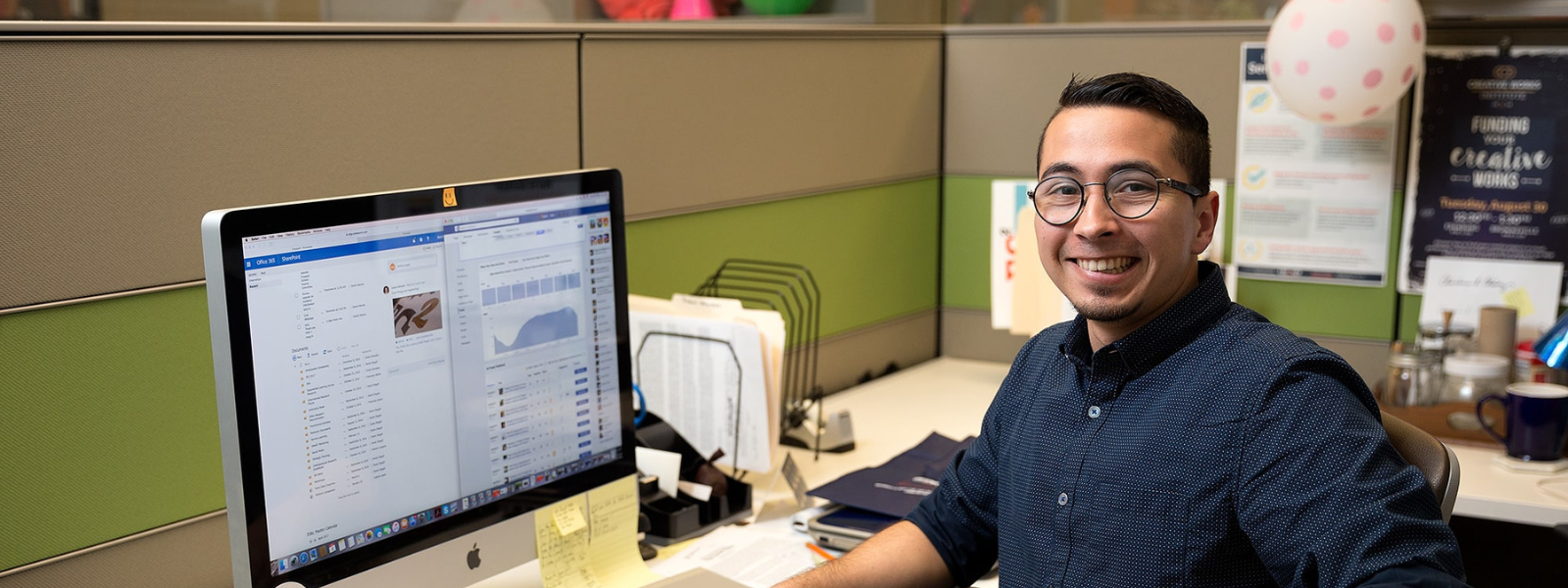 Employee in front of computer inside cubicle smiles at the camera. 